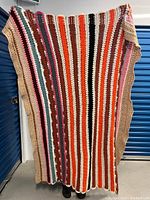 Full view of a colorful crochet blanket with vertical stripes in various colors including orange, red, brown, green, pink, and beige tones.