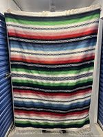 Full view of colourful woven blanket or rug with horizontal stripes in green, red, white, and black, held vertically for size reference.