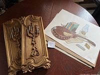 Photo showing a decorative gold-tone wall sconce with two candlestick holders next to framed art prints depicting musical instruments and vintage objects on a wooden table.