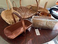 Seven assorted baskets on a wooden table including wicker and wooden styles, one wrapped in plastic labeled Longaberger Vanity Tray.