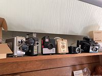 Wide shot showing six vintage cameras lined up on a wood shelf with a cardboard box of accessories to the left.