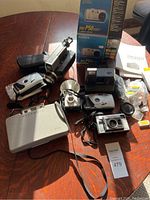 Photo showing vintage cameras and accessories on table with natural light highlighting various devices including Polaroid 320, Sony camcorder, and compact cameras.