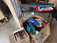 Wide view including black folding table, box of clothing, box of books, and tent box, illustrating entire lot contents.