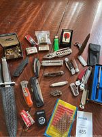 Full view of all items spread on wooden surface including knives, lighters, coin, cufflinks, pens, ammunition, and accessories.
