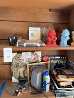 Shelf displaying various decor items, a framed memo board, colorful small statues, books stacked and arranged, and electronics including a modem and compressed air can.