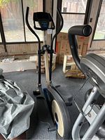 Elliptical trainer with black handlebars, digital display, and beige blue body near windows.