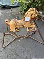 Side view of molded horse figure on metal frame base placed outdoors on pavement with surrounding foliage and garage area in background