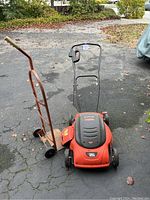 Black & Decker electric lawn mower and rusty metal hand truck side by side on asphalt.