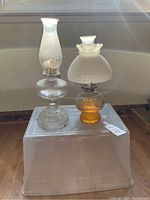 Clear and amber glass vintage oil lamps placed side by side on a plastic box in front of a window with sunlight shining.
