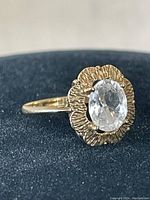 Close-up front-view of 10 kt gold ring with clear oval faceted stone in floral bezel setting on dark background.