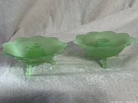 Pair of green Fenton jadeite glass candle holders with scalloped flower-like rims and three legs on light fabric background