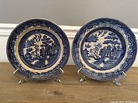 Pair of antique Spode Blue Willow pieces: one plate and one bowl, placed on stands on wooden surface against a white baseboard background. Both show characteristic Blue Willow oriental design in blue and white.