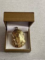 Gold-toned Jesus pendant displayed inside a brown jewelry box with white satin lining.