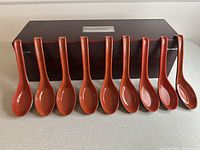 Nine reddish-brown lacquered Chinese spoons lined up in front of a dark wood storage box with a silver plaque.