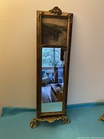 Front view of the tall antique mirror showing the black and white print at top and clear mirror below, ornate gold frame with damage to top edge.