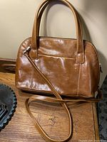 Brown leather handbag with top handles and a detachable long shoulder strap, front view on a wooden surface.