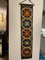 Full view of the vintage needle point bell pull wall hanging showing repeated geometric flower motifs in orange, blue, green, and brown colors on a dark background. Measures approximately 41" H x 8.75" W.