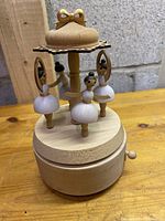 Front view of the wood musical box with 3 attached ballerina figures in white skirts and black hair, wooden bow decoration on top, and winding key at base.