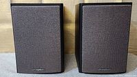 Pair of iSymphony M1 bookshelf speakers with black fabric covers on, showing front view.