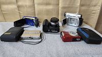Lot of five digital cameras and camcorders shown from front angle on gray fabric surface, various models and colors visible including DSLR, compact cameras, and camcorders.