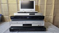 Stacked electronic players including portable DVD player on top, Sony Blu-ray, Yamaha player, and DVD/VHS combo units beneath