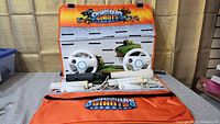 Lot contents showing 2 white Wii steering wheels, 2 white Wii Remote controllers with Motion Plus, 2 white Wii Nunchucks, and part of the orange Skylanders Giants wall hanging storage board behind them