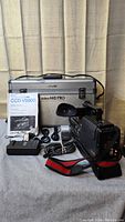 Full view of Sony CCD-V5000 Hi8 video camera with hand strap, manual, cables, charger, batteries, and silver hardshell carrying case with black handle.