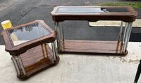 Two tables outdoors on a concrete surface showing wood surfaces, glass tops, and metal corner columns.