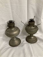 Two tarnished metal oil lamp bases side by side against a white background showing overall shape and height.