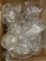 Top view of assorted clear glass and crystal glasses, some with cut patterns and others plain, arranged tightly in a box.