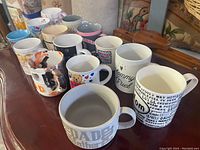 14 assorted coffee mugs on wooden table, various designs and colors, one chipped mug visible