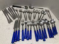 Photo showing assorted stainless steel silverware including knives, forks, and spoons with blue plastic handles and plain stainless steel handles arranged on white background.