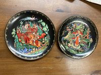 Two decorative Bradford Exchange plates on wooden surface showing artwork and gold leaf edging.