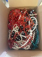 Photo of tangled costume jewelry necklaces in a cardboard box showing multiple colors of beads including red, white, black, cream, and turquoise.