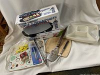 Photo showing George Foreman grill box, Pampered Chef covered divided dish, spatula, tongs, cutting board, serving tray, and assorted utensils including grill brush, skewers, and corn holders.