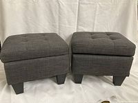 Both gray upholstered storage ottomans shown side by side with closed tufted tops and plastic legs visible.