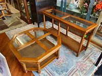 Two wooden tables; one rectangular console or sofa table with a glass top and caned lower shelf, and one octagonal coffee table with glass top and caned lower shelf, placed on a rug in a room with wooden flooring.