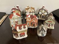 Eight miniature Christmas village buildings arranged on a dark surface, showing various styles including houses, church, and school with snow detail on roofs.