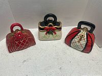 Three handbag-shaped ceramic jars with different designs: red with gold floral, cream with red bow, and plaid with gold lock, shown on white table.