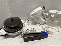 Four small kitchen appliances on a white surface: Black & Decker hand mixer with beaters, Regal electric slicer secured with blue tape, egg cooker with dome lid, and Sunbeam can opener.