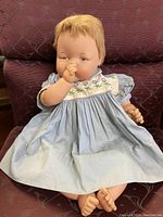 Full doll seated with light blue dress, cloth body, light brown hair, and hand near mouth.