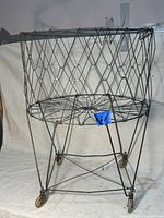 Full front view of the antique folding metal laundry basket showing wire lattice sides, metal frame, and caster wheels. The basket is standing upright in an open, unfolded position.