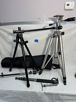 Two camera tripods, one in silver and one in black, with extension arm and tripod accessories laid out on a white surface.