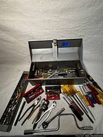 Open metal toolbox showing various hand tools spread out including screwdrivers, pliers, wrenches, and ratchets.