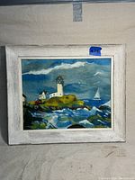 Frontal view of the framed oil painting showing the lighthouse, rocky coastline, ocean waves, and sailboat under a cloudy sky.