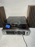 Photo showing the main stereo unit with the built-in turntable under a closed clear plastic dust cover, two wooden speakers on either side.
