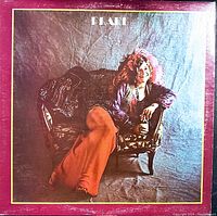 Photo of the front cover of the Janis Joplin 'Pearl' vinyl record showing Janis Joplin seated on a chair with vibrant clothing and feathered headpiece. Some wear visible on cover edges and surface.