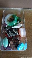 Assorted polished stones and glass crystal pendants displayed inside a clear container on a flat surface.
