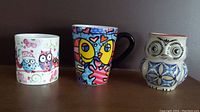 Three mugs shown on a shelf: colorful duo owl patterned mug, Romero Britto fish mug, and 3D embossed owl mug with blue, black and brown details and coral rim.