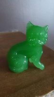 Green translucent jade glass cat figurine shown from a side angle on a wooden surface with visible stylized swirl pattern on tail and body.